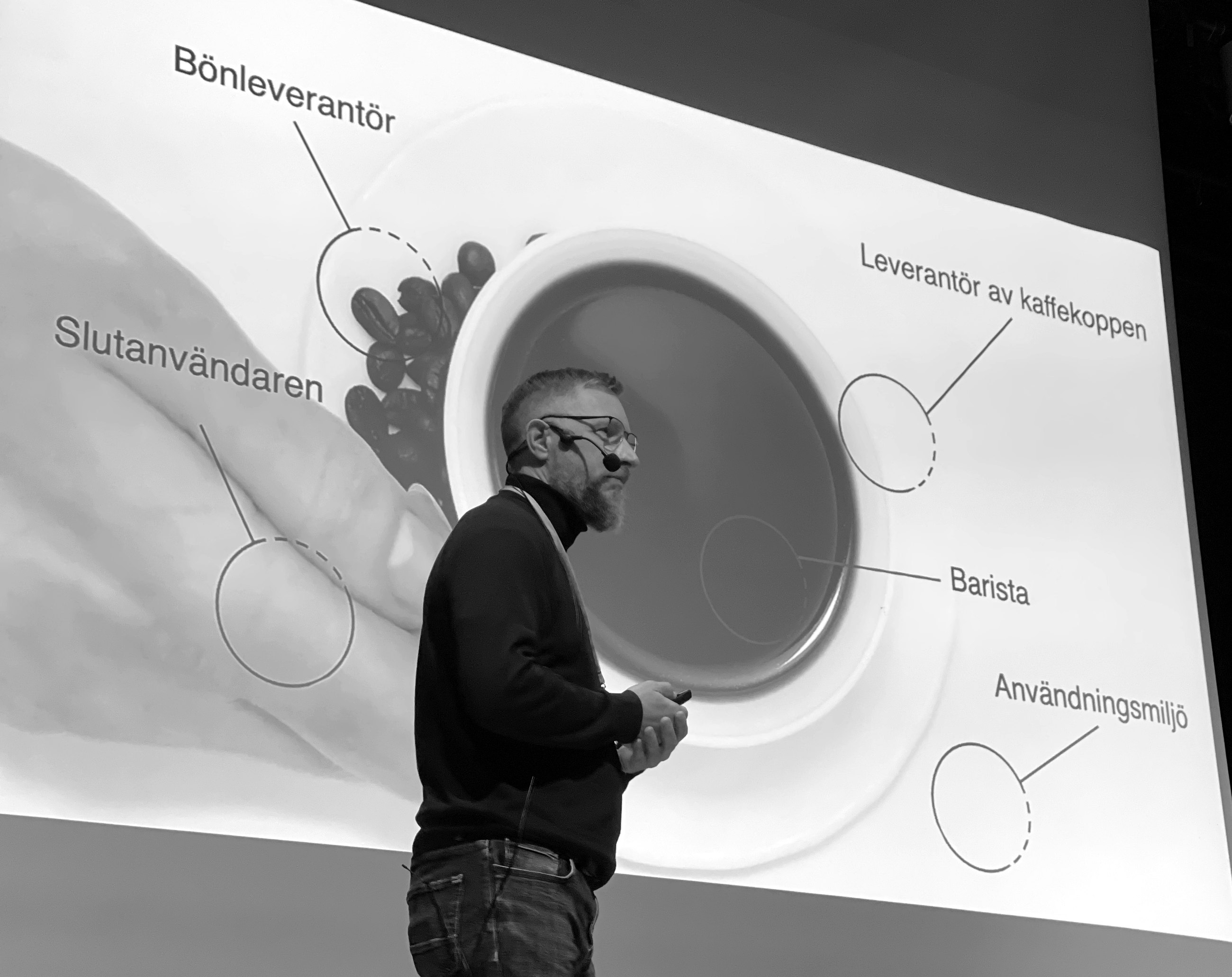 Mathias Duell på scen framför en projicerad kaffekopp där varje aktör i värdenätverket är utpekad: bönleverantör, barista, leverantör av kaffekoppen, användningsmiljö och slutanvändaren